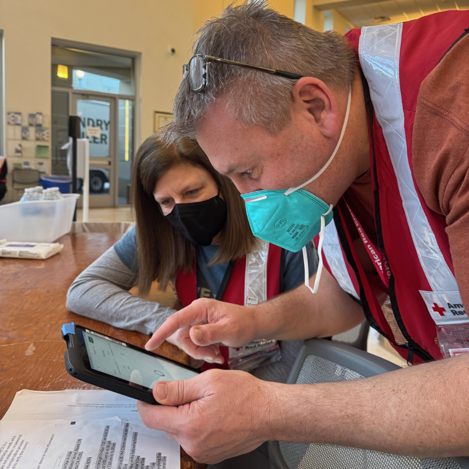 Two people in Red Cross vests and masks look at a tablet in a shelter. 