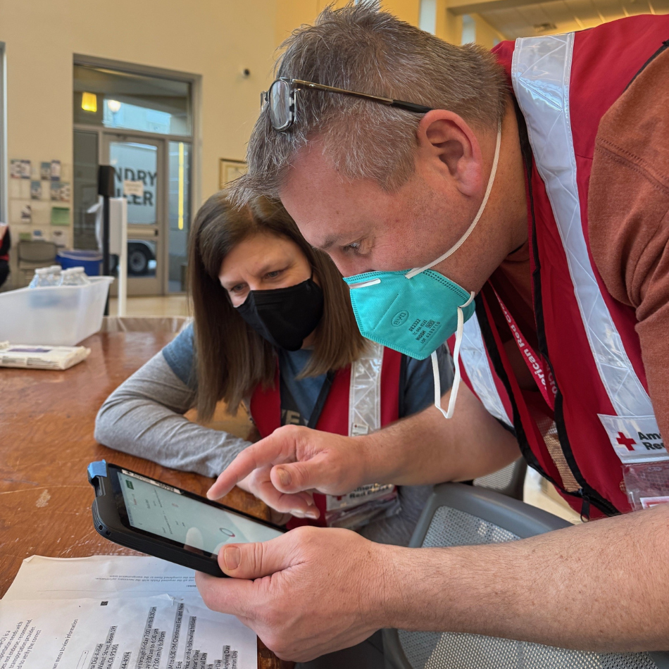 Two people in Red Cross vests and masks look at a tablet in a shelter.