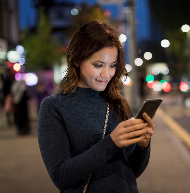 A woman smiles at her smartphone on a city street at night. 