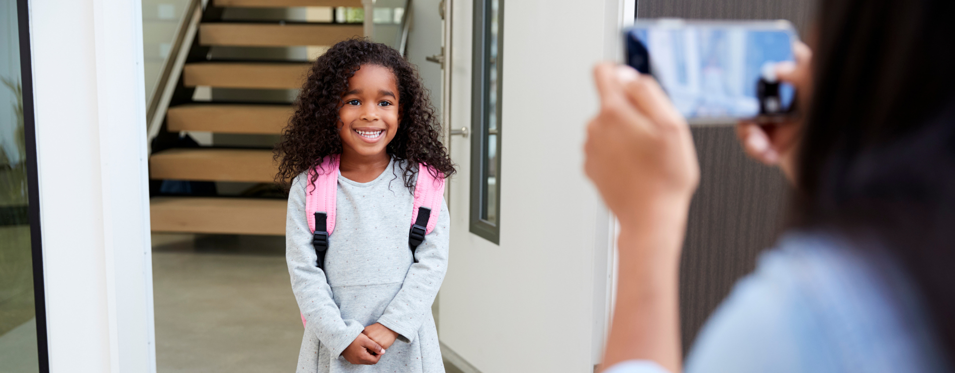 A young girl wearing a backpack poses for for her first day of school photo for her mother in her doorway.