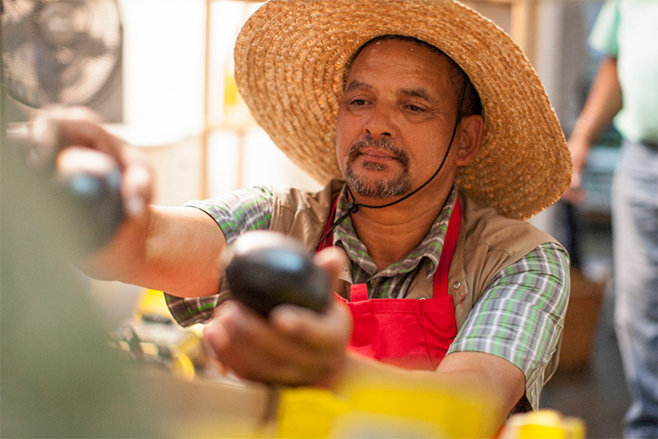 A man in a straw hat and aprons arranges vegetables at a farm stand.