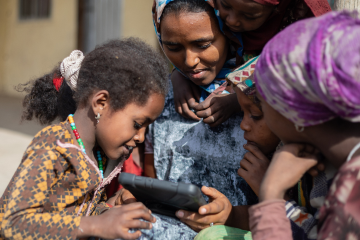 Several women and a young girl look at a tablet. 