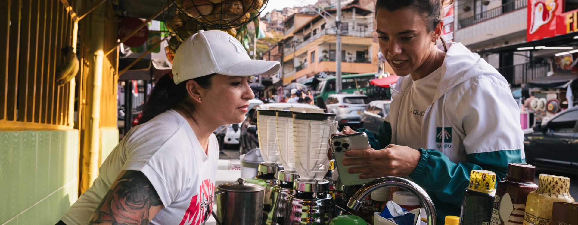 A woman wearing a white baseball hat speaks with another woman holding a smartphone. 