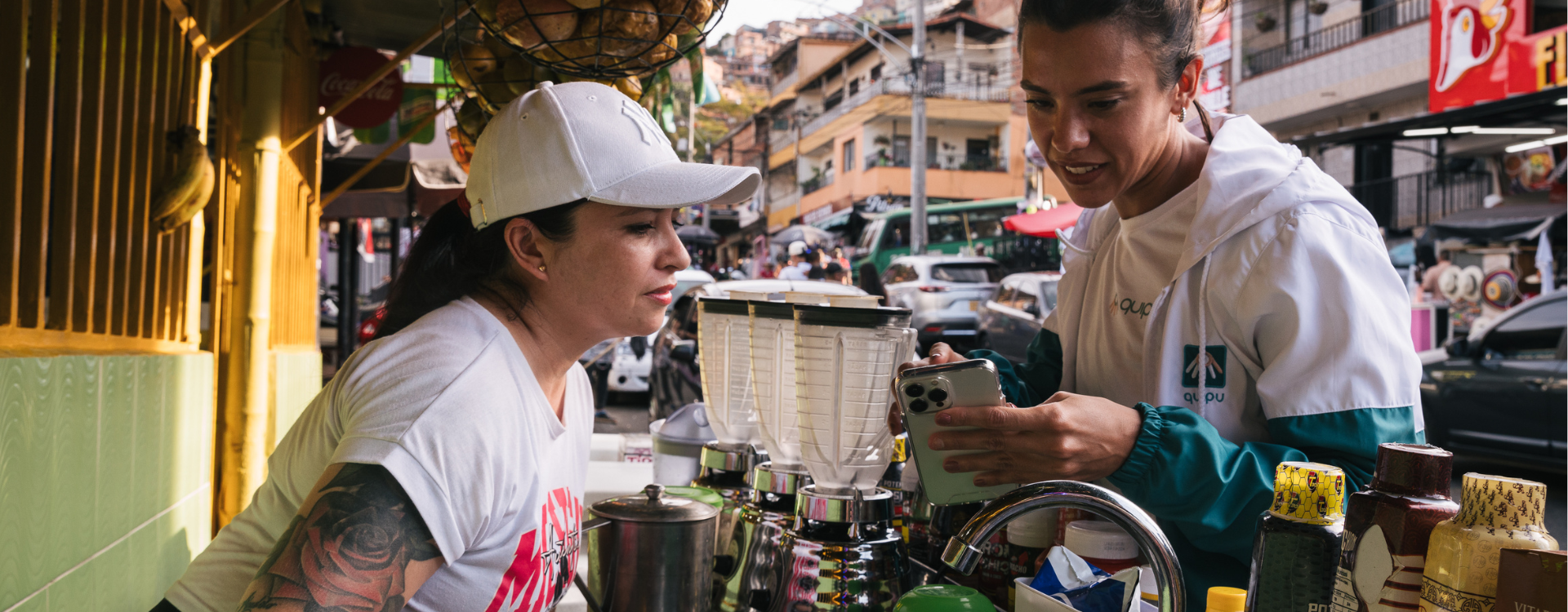 A woman wearing a white baseball hat speaks with another woman holding a smartphone.
