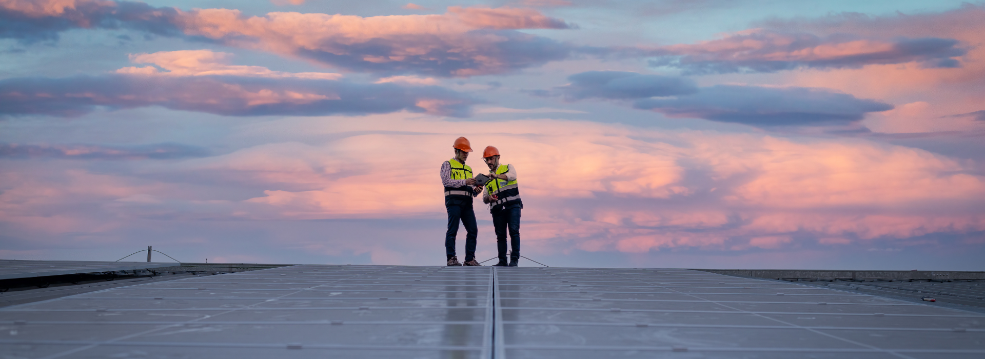 Two men in work vests and hard hats look at a tablet while standing on a field of solar panels at sunrise.