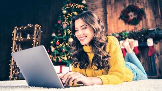 A woman in a yellow sweater lies on the floor in front of a Christmas tree looking at her laptop. 