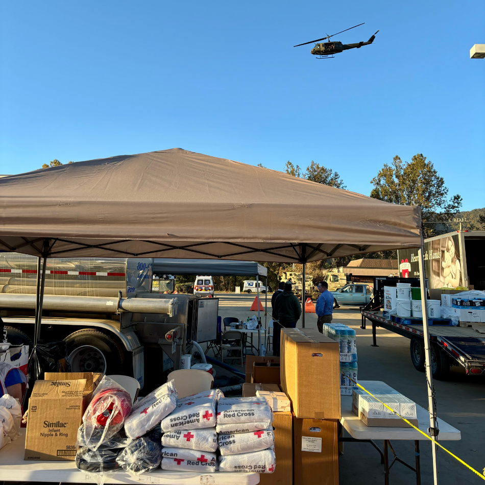 A tent set up at a Red Cross community care center with supplies for Hurricane Helene victims. A helicopter flies overhead.