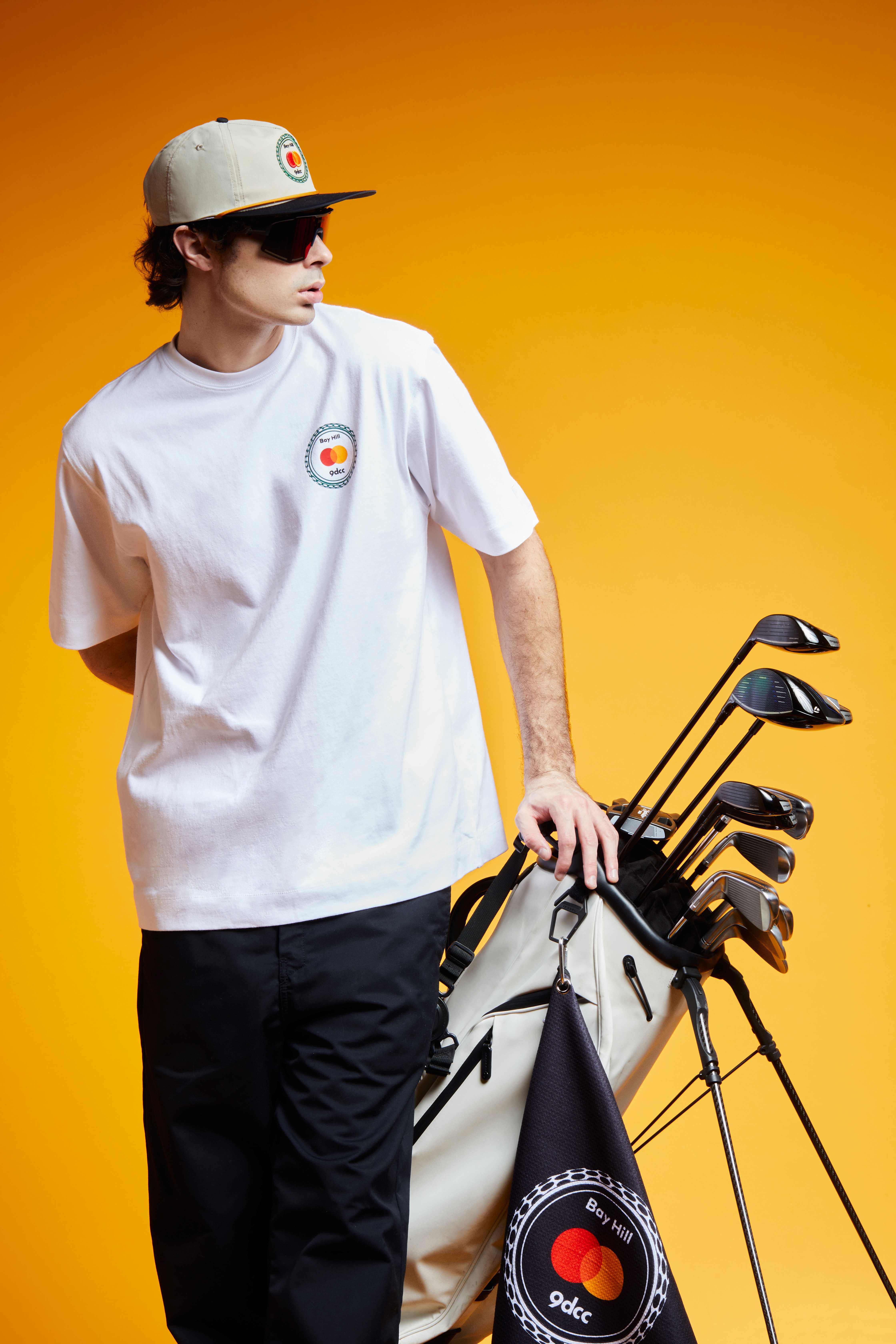A man wears a Mastercard and 9dcc-branded T-shirt and hat next to golf clubs.