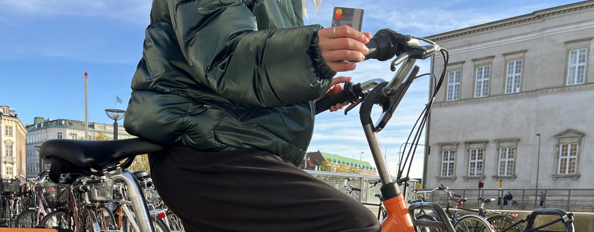 A woman in a puffer jacket sits on a bicycle and holds a Mastercard. 