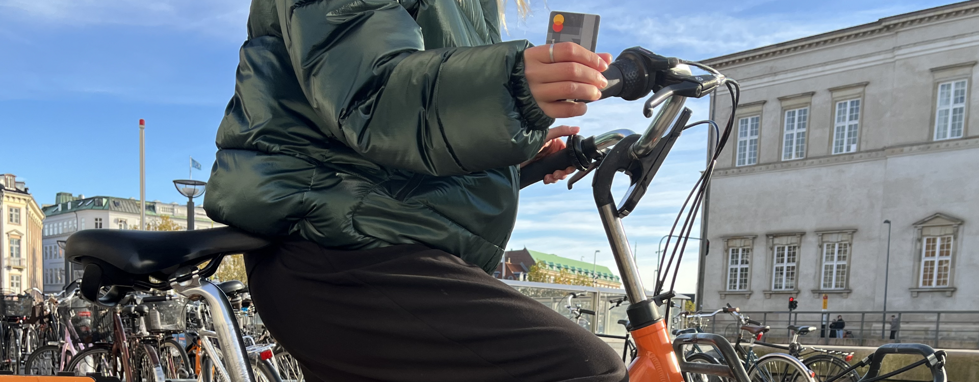 A woman in a puffer jacket sits on a bicycle and holds a Mastercard.
