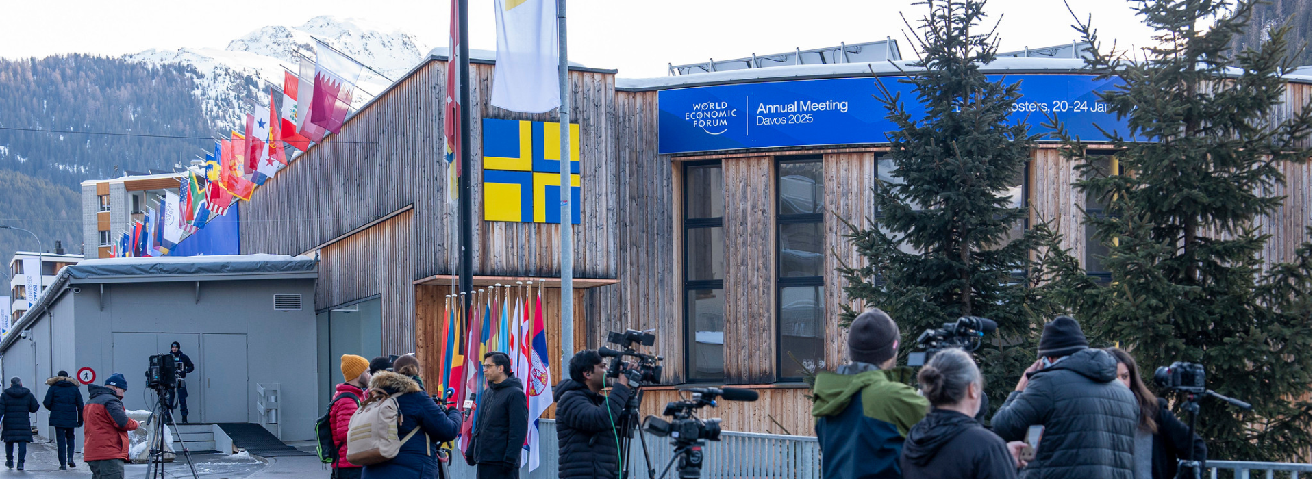 The exterior of a building where Davos 2025 is being held, with flags along the roofline and camerapeople setting up in front. 