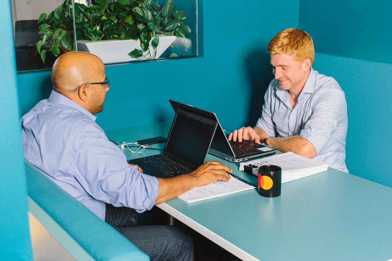 Two co-workers sit at a table with their laptops.